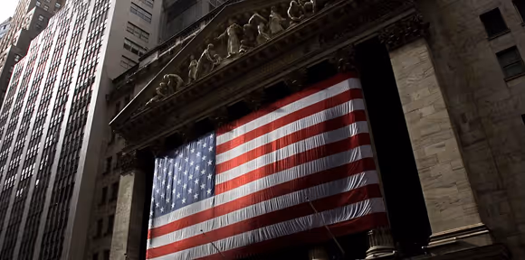 Large American flag hanging on the facade of a historic building in a city with tall skyscrapers.